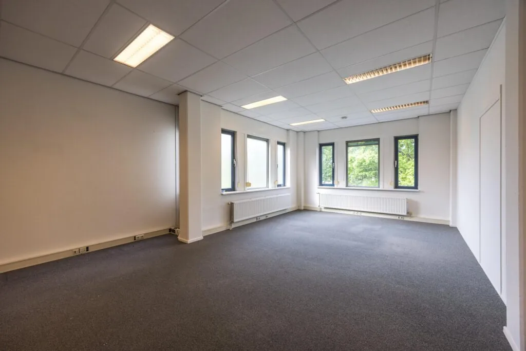 Lichte, lege kantoorruimte aan de Buitenweg met grijze vloerbedekking en meerdere ramen.