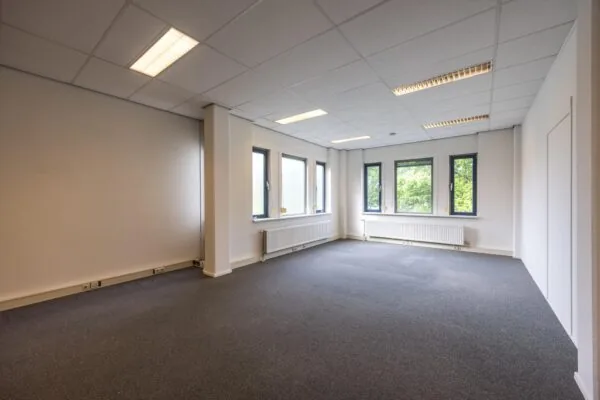 Lichte, lege kantoorruimte aan de Buitenweg met grijze vloerbedekking en meerdere ramen.
