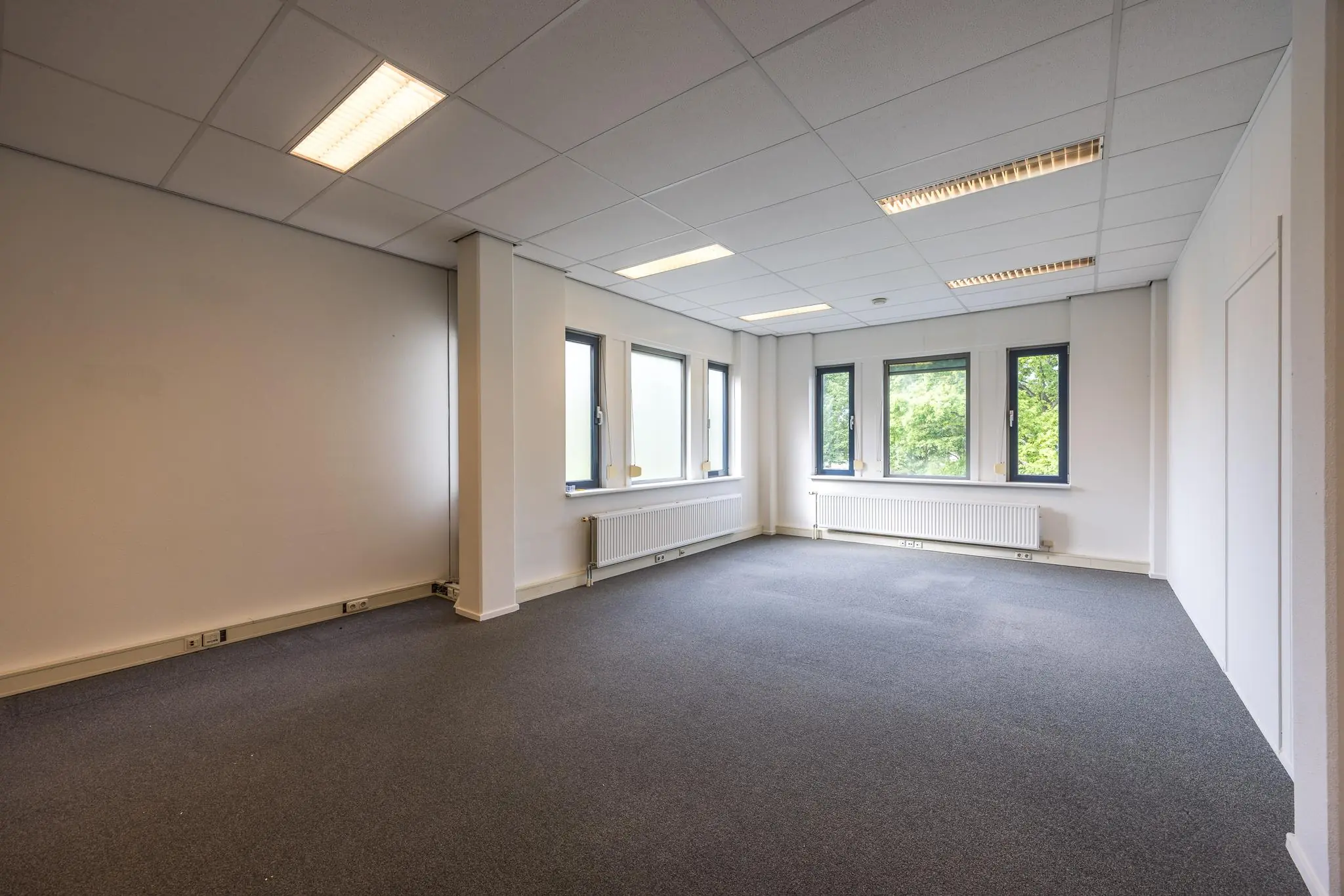Lichte, lege kantoorruimte aan de Buitenweg met grijze vloerbedekking en meerdere ramen.