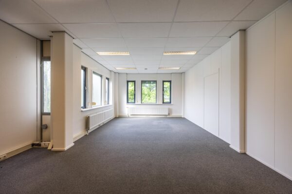 Leeg kantoor met grijze vloerbedekking, witte muren en meerdere ramen aan de buitenwegzijde.