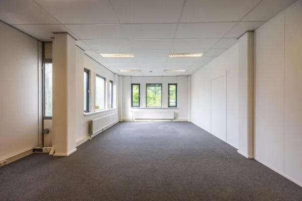 Leeg kantoor met grijze vloerbedekking, witte muren en meerdere ramen aan de buitenwegzijde.