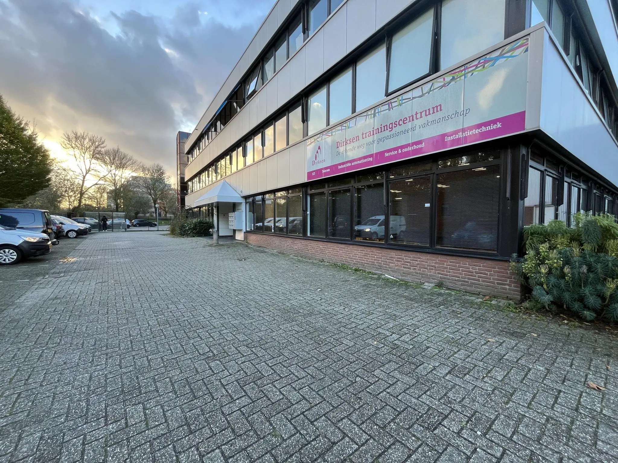 Vooraanzicht van het Dirksen trainingscentrum aan de Industrieweg met parkeerplaatsen en een ondergaande zon op de achtergrond.