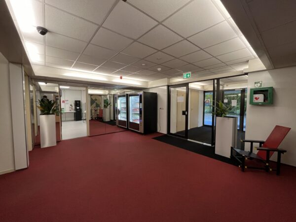 Hal met rode vloerbedekking, frisdrankautomaten, plantenbakken, een rode designstoel en een AED-kast aan de muur.