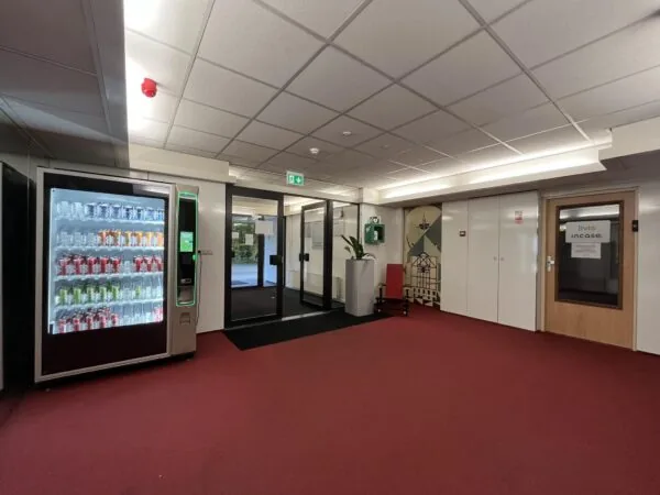 Hal met rode vloerbedekking, frisdrankautomaat, glazen deuren en een deur met het bordje "Livis incase".