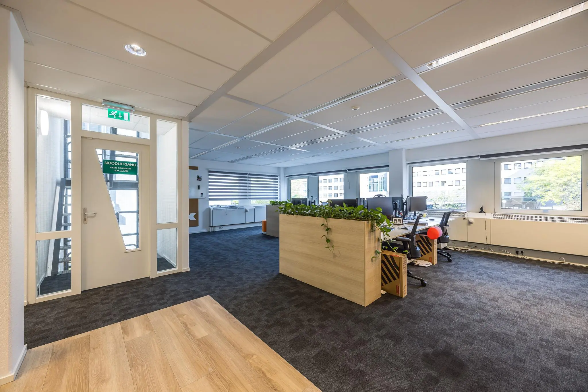 Moderne kantoorruimte aan de Varrolaan met werkplekken, plantenbak en zicht op de nooduitgang.