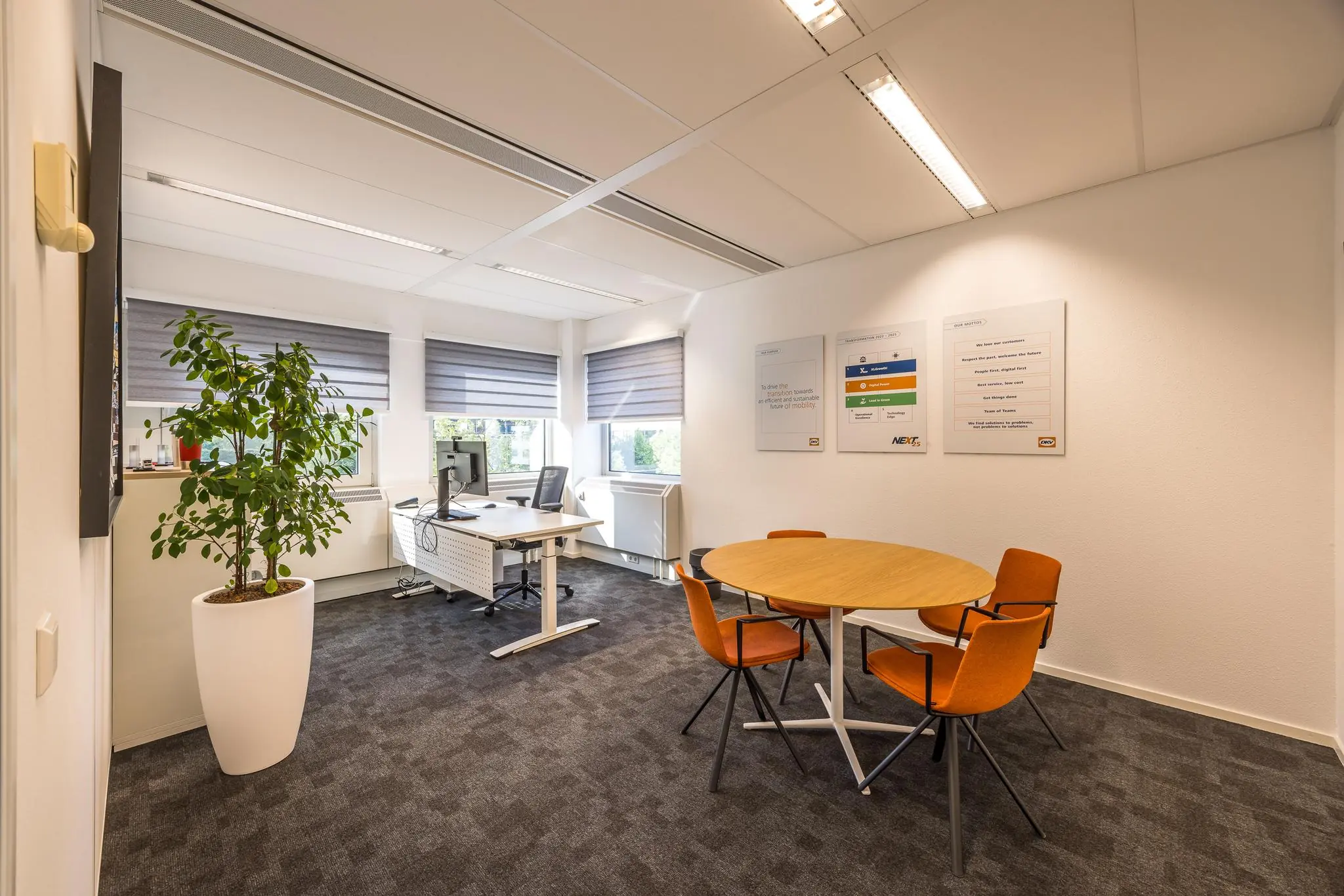 Modern kantoorinterieur met een bureau, vergadertafel met vier oranje stoelen, een grote plant en informatieve posters aan de muur.