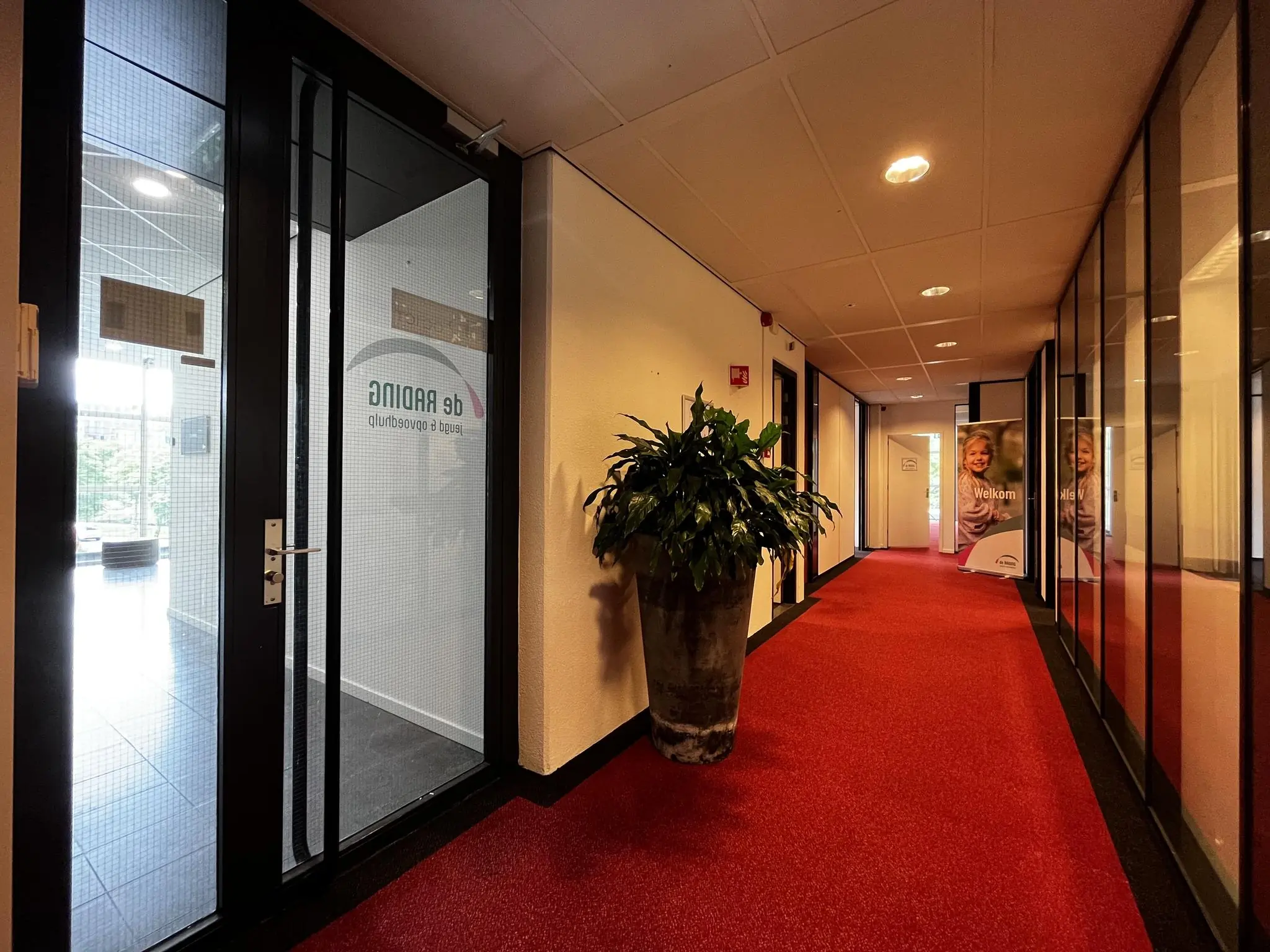 Gang in een kantoorpand aan de Vreeswijksestraatweg met rode vloerbedekking, glazen deuren en een banner van jeugdzorgorganisatie De Rading.