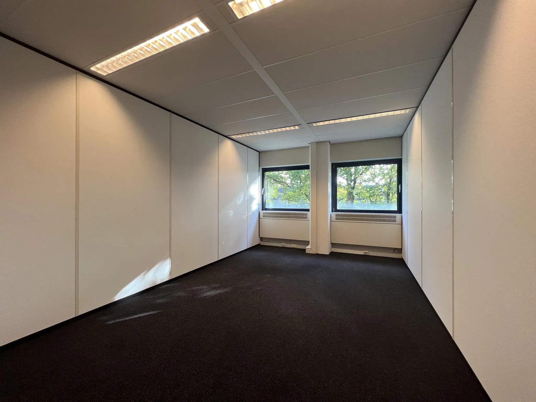 Lege kantoorruimte aan de Vreeswijksestraatweg met witgelakte wanden, zwart tapijt en drie ramen met uitzicht op bomen.