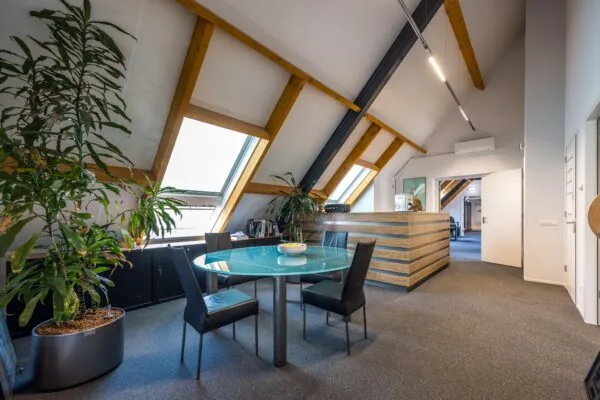 Moderne kantoorruimte met glazen vergadertafel, planten en houten receptiebalie onder een schuin dak met dakramen.