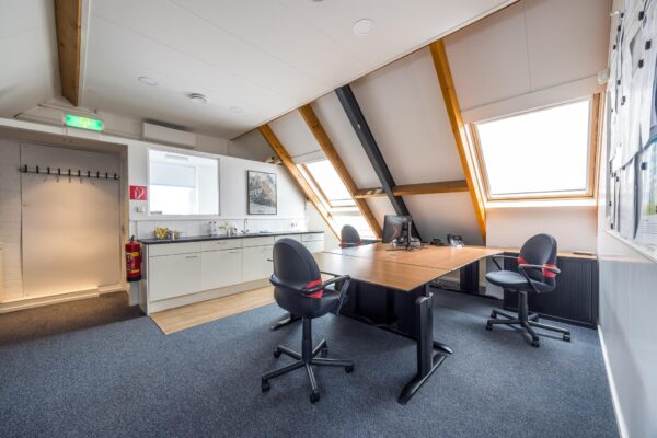 Modern kantoor op zolderverdieping met schuine daken, werkplekken, keukenblok en veel daglicht.