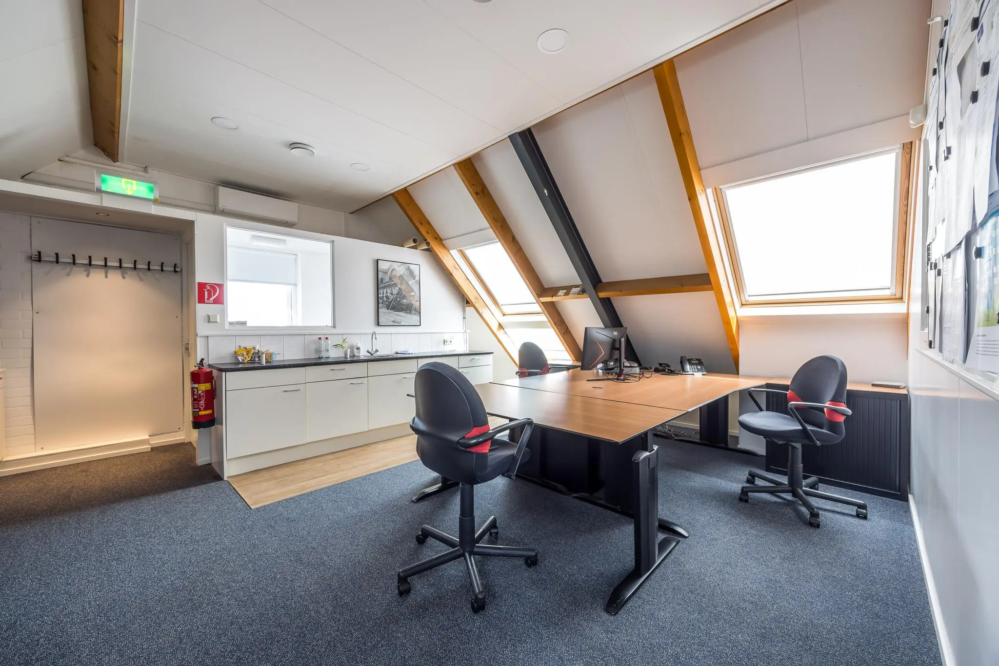 Modern kantoor op zolderverdieping met schuine daken, werkplekken, keukenblok en veel daglicht.