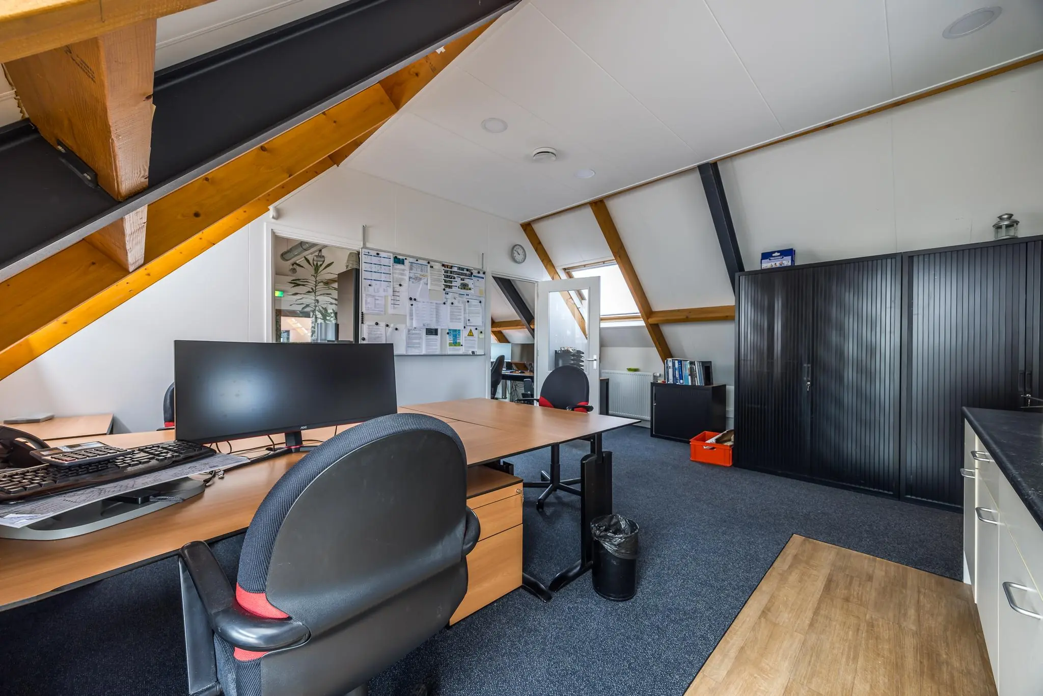 Modern kantoor met schuine houten dakbalken, meerdere bureaus, een prikbord en zwarte kasten.