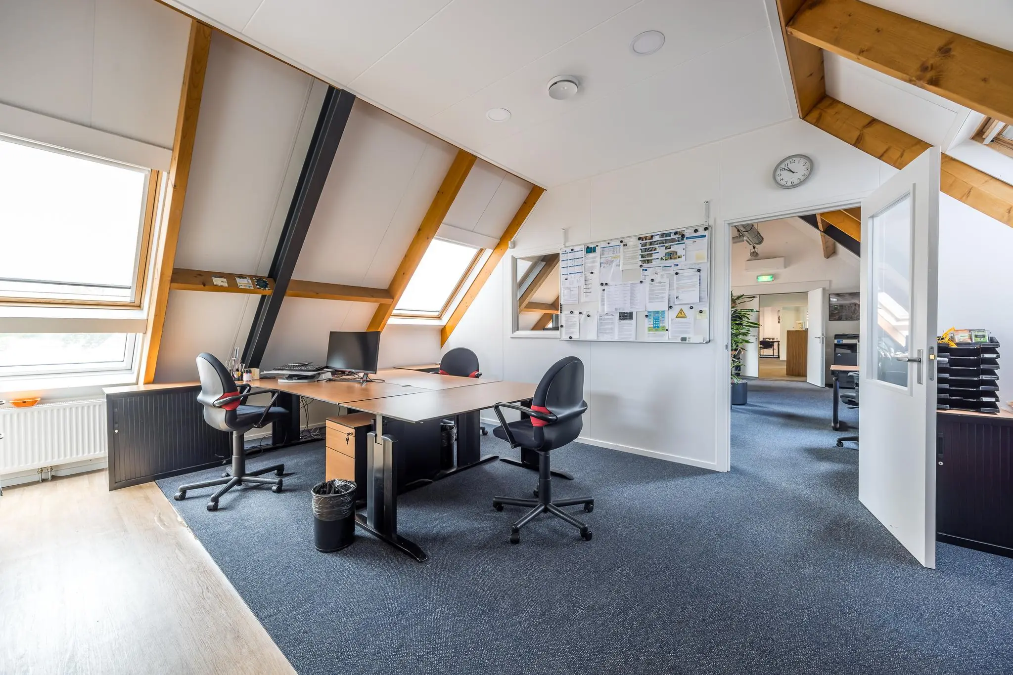 Kantoorruimte op de Voorveste met houten balken, schuine dakramen, bureaus, stoelen en een prikbord aan de muur.
