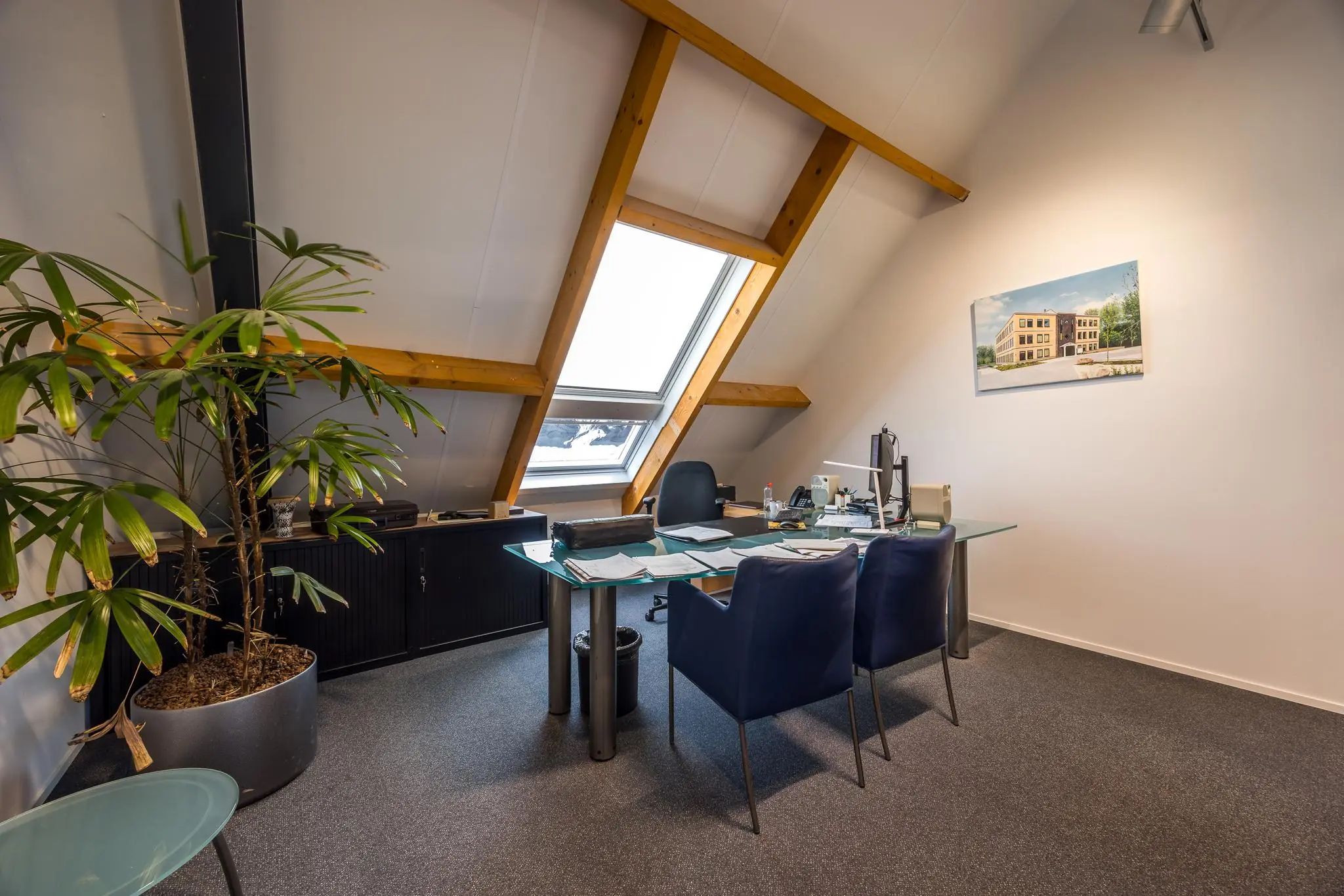 Modern kantoor op zolderverdieping met glazen bureau, dakraam en een schilderij van gebouw Voorveste aan de muur.