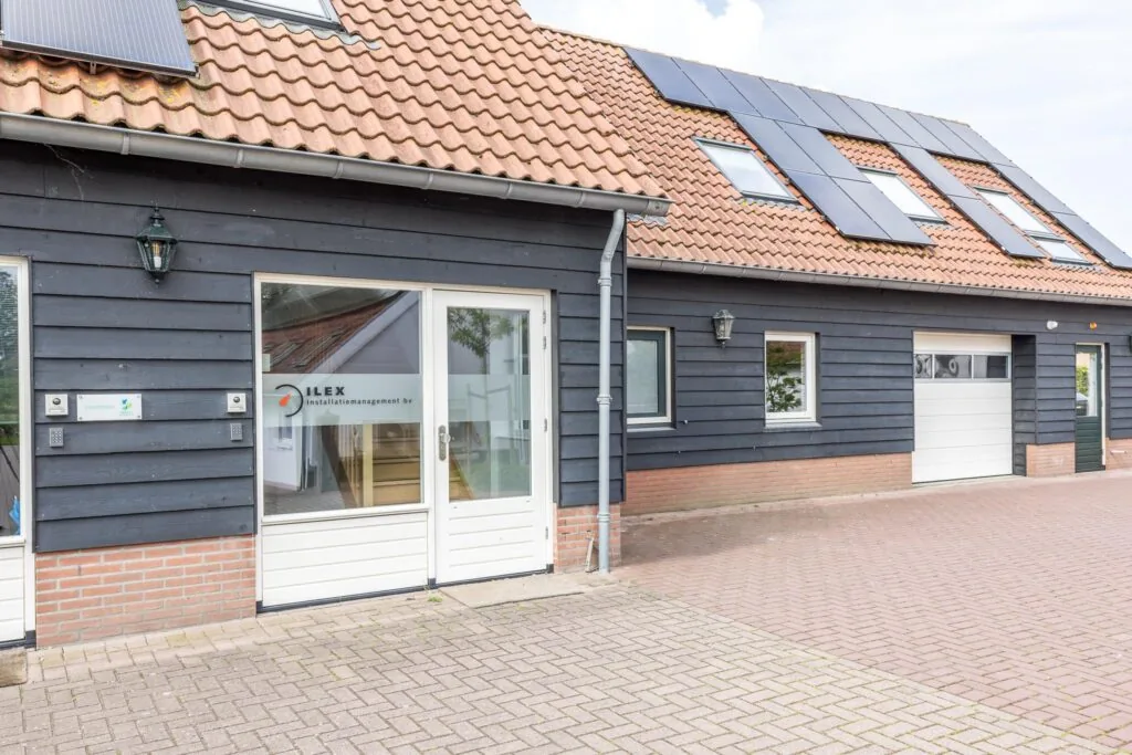 Vooraanzicht van het bedrijfspand van ILEX Installatiemanagement BV met zwarte houten gevel, zonnepanelen op het dak en een witte garagedeur.