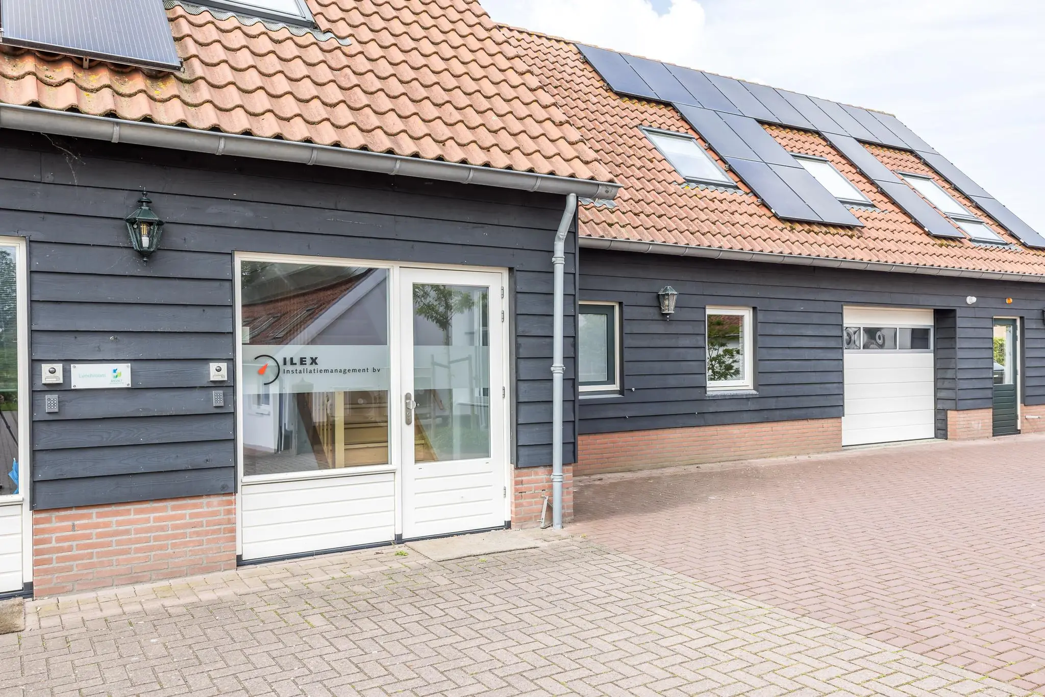 Vooraanzicht van het bedrijfspand van ILEX Installatiemanagement BV met zwarte houten gevel, zonnepanelen op het dak en een witte garagedeur.