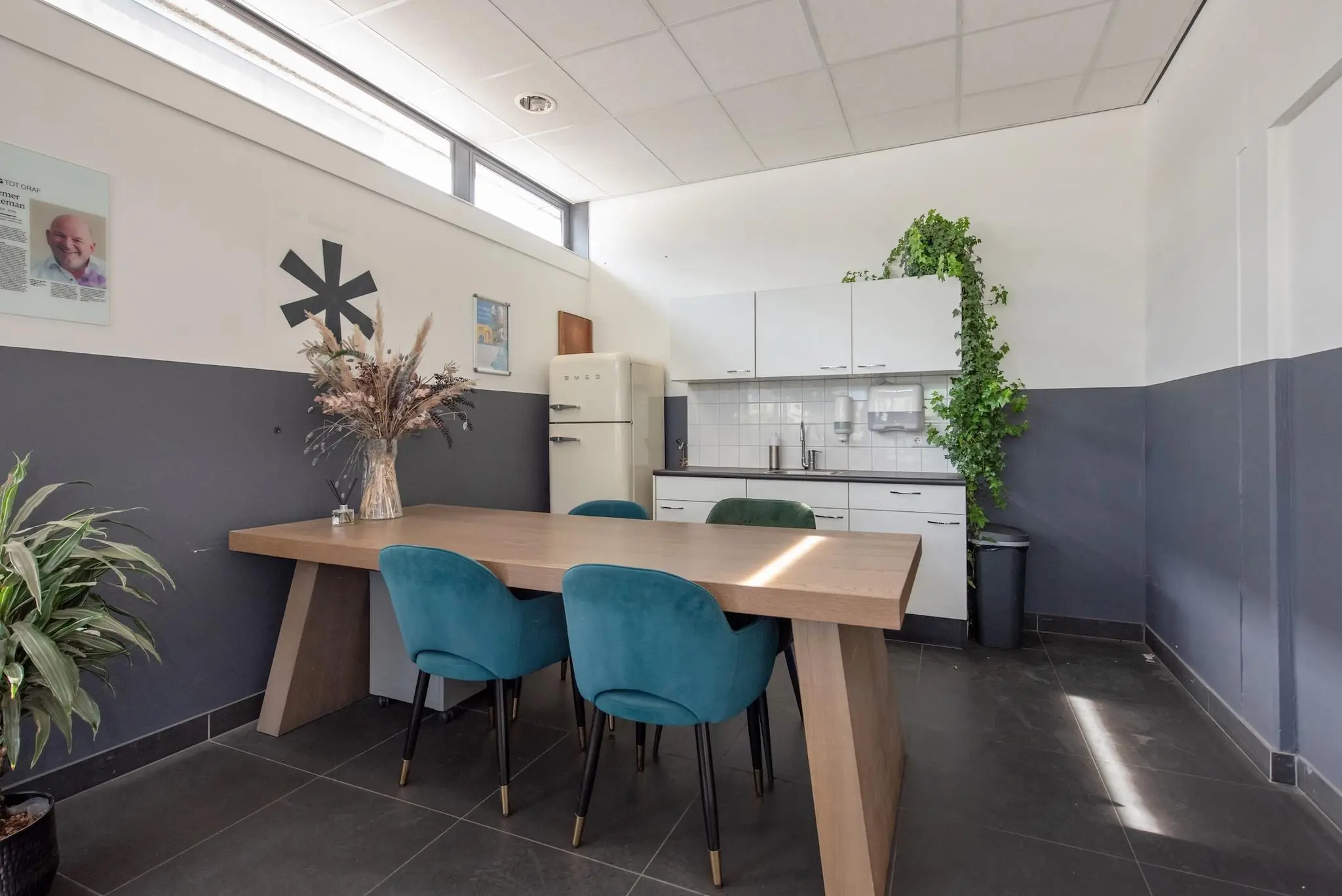 Moderne kantine aan de Limpergstraat met een houten tafel, blauwe stoelen, witte keuken en planten.