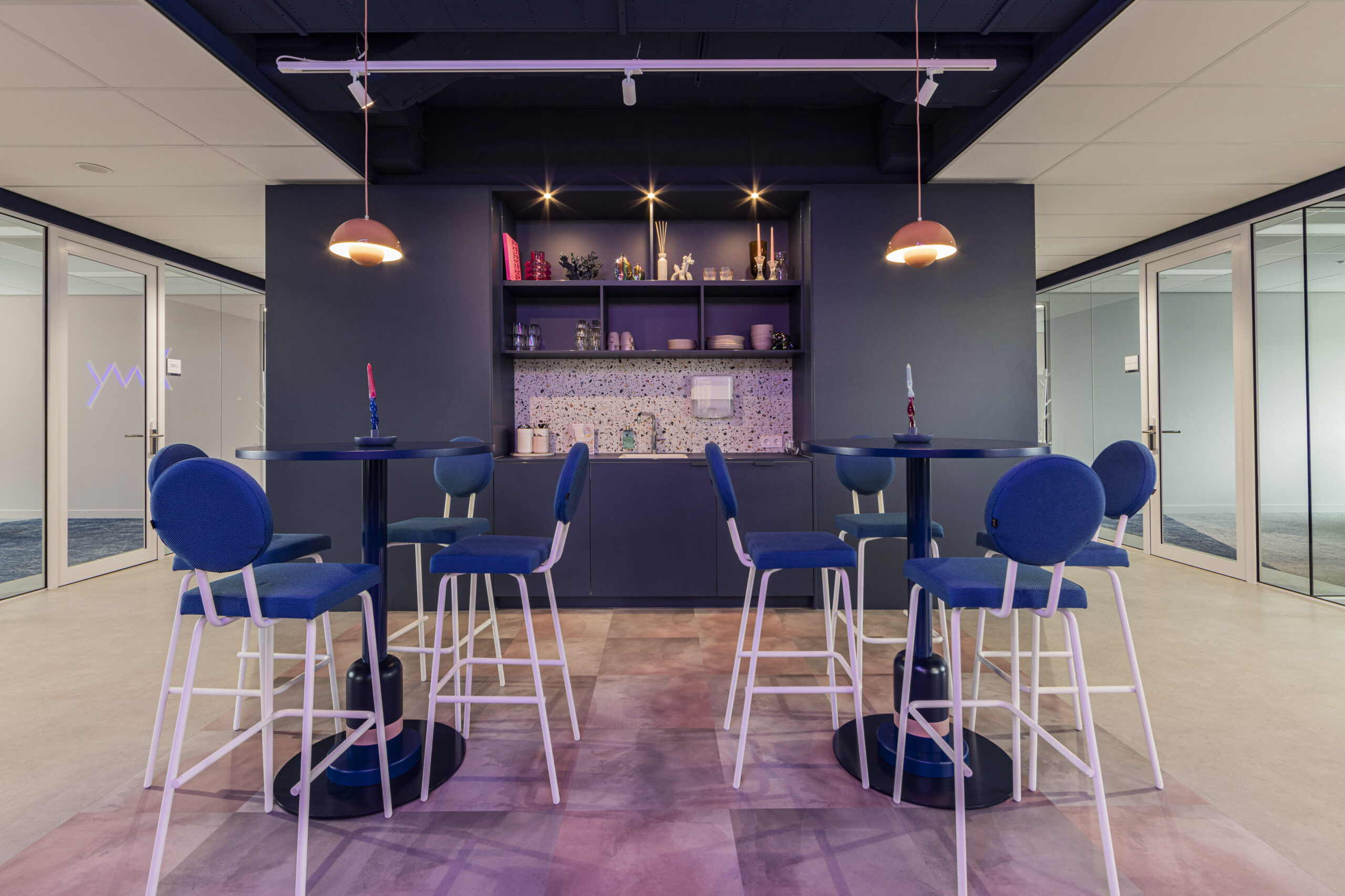 Een modern kantoor met een kitchenette en hoge tafels met blauwe stoelen.