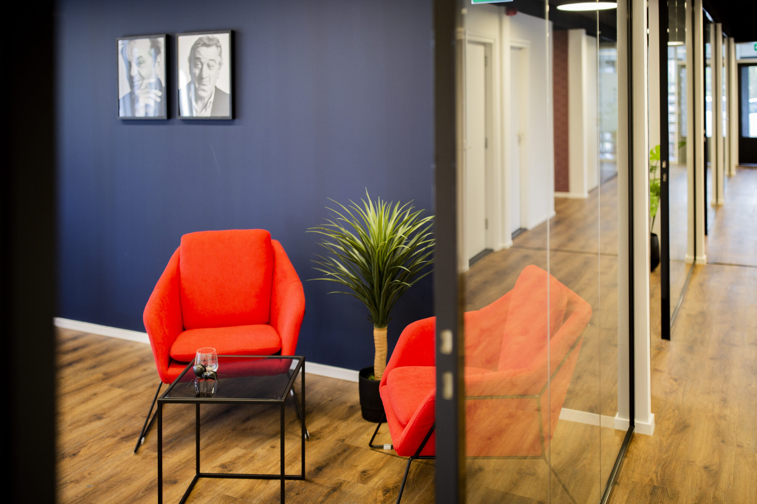 Een moderne kantoorruimte met een feloranje stoel, glazen bijzettafel en groene plant naast een glazen wand.