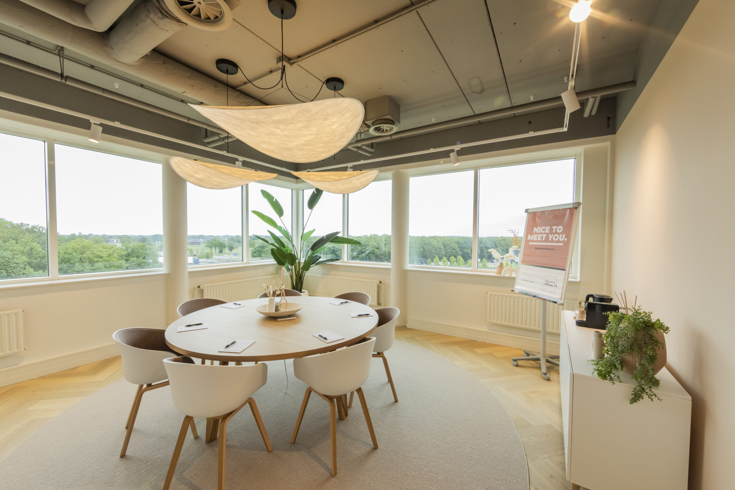 Een moderne vergaderruimte met een ronde tafel, stoelen, grote ramen en planten.