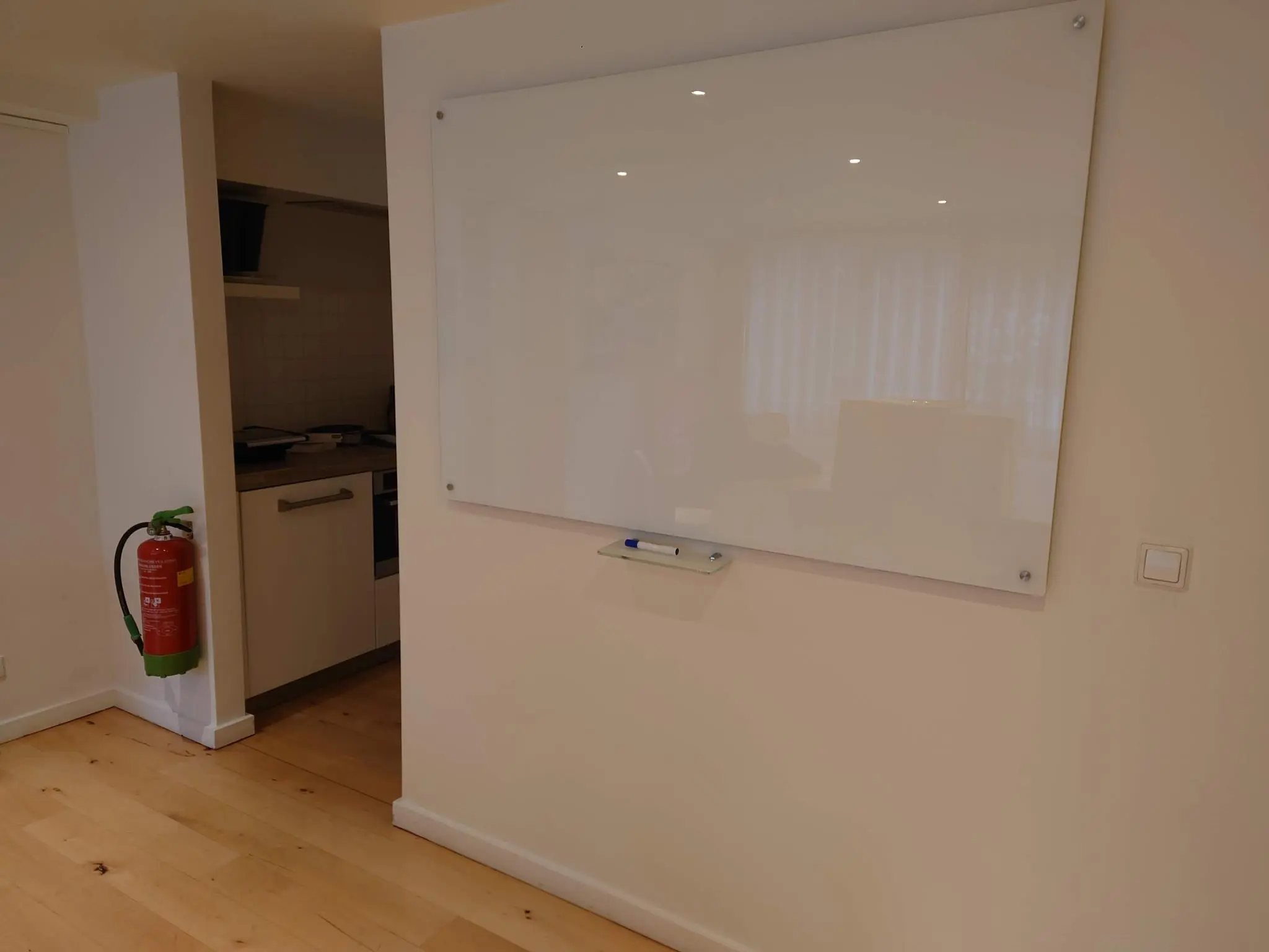 Witte glazen wandwhiteboard aan de muur in een lichte ruimte naast een open keuken en een brandblusser.