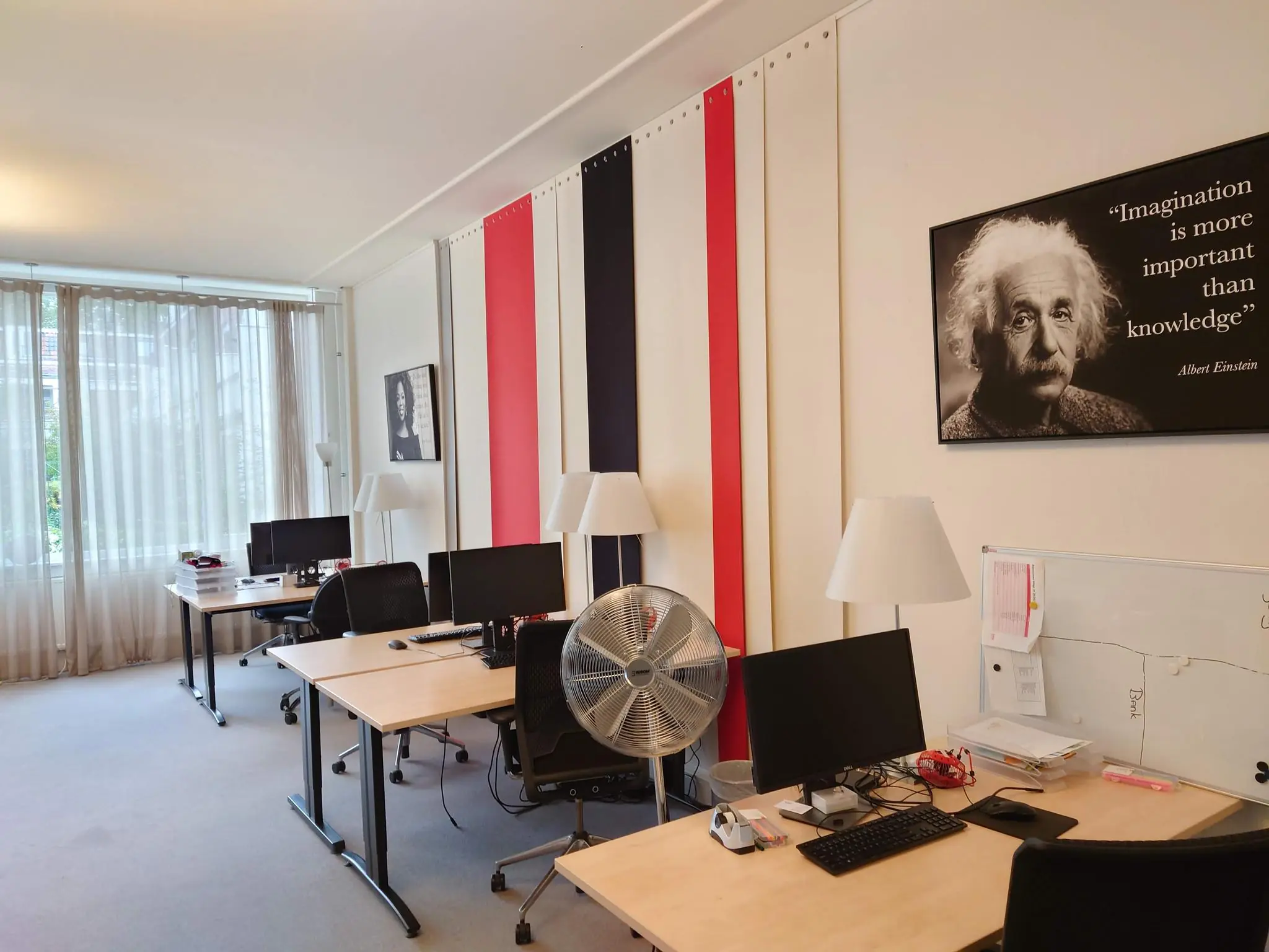 Modern kantoor met meerdere werkplekken, computers, een grote ventilator en een muurposter van Albert Einstein met het citaat "Imagination is more important than knowledge".
