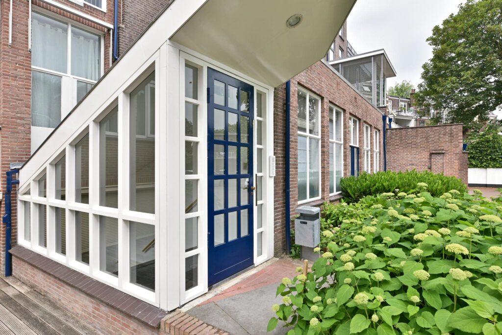 Ingang van een bakstenen gebouw aan de Maliesingel met blauwe deur, glazen entree en groene planten ervoor.
