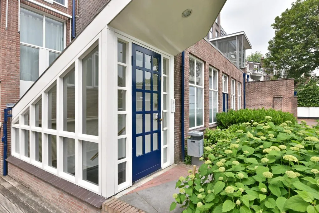 Ingang van een bakstenen gebouw aan de Maliesingel met blauwe deur, glazen entree en groene planten ervoor.