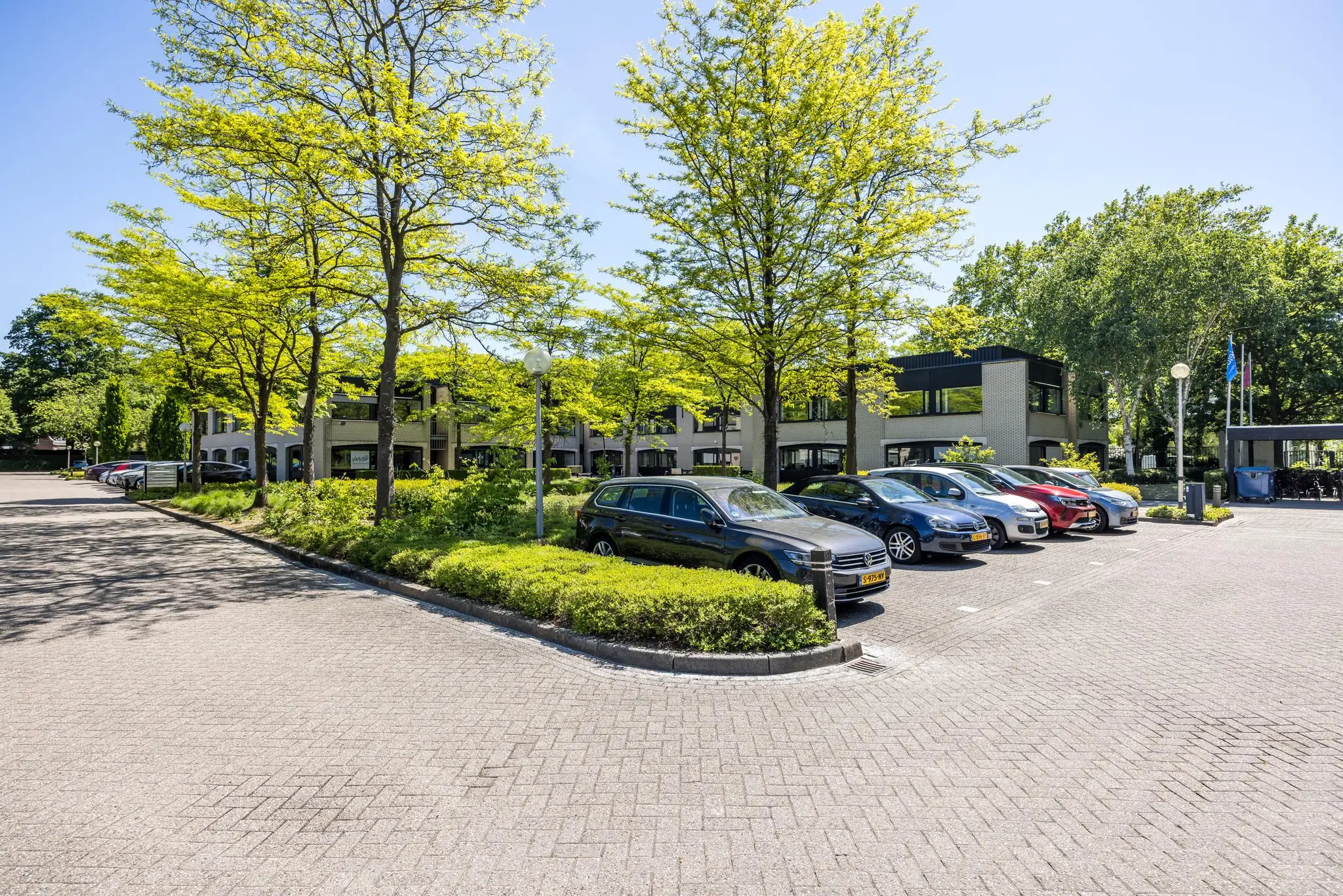 Parkeerterrein met geparkeerde auto's en groene bomen bij een modern gebouw aan de Burgemeester Verderlaan.