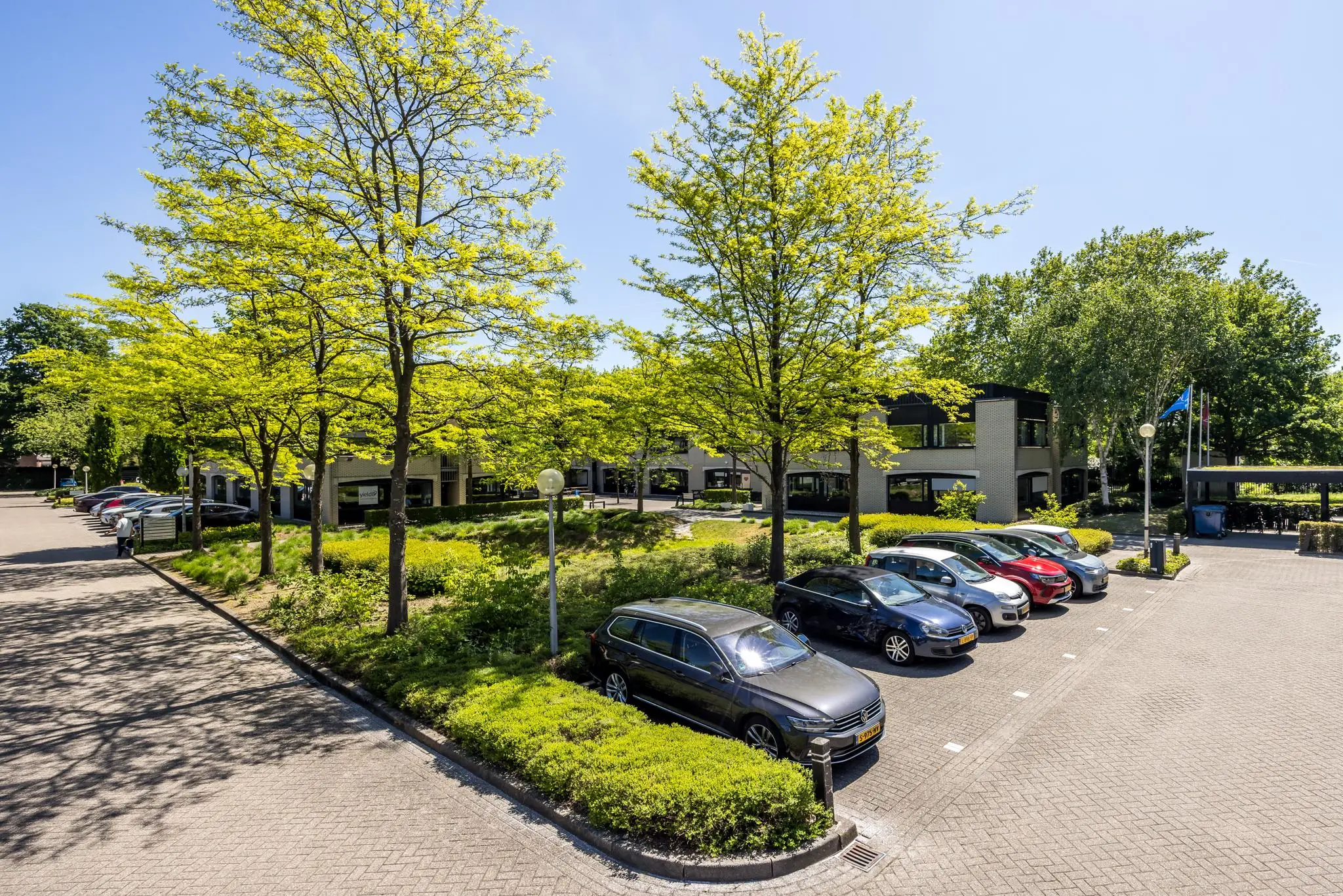 Parkeerterrein met geparkeerde auto's en een modern kantoorgebouw aan de Burgemeester Verderlaan, omgeven door groene bomen.