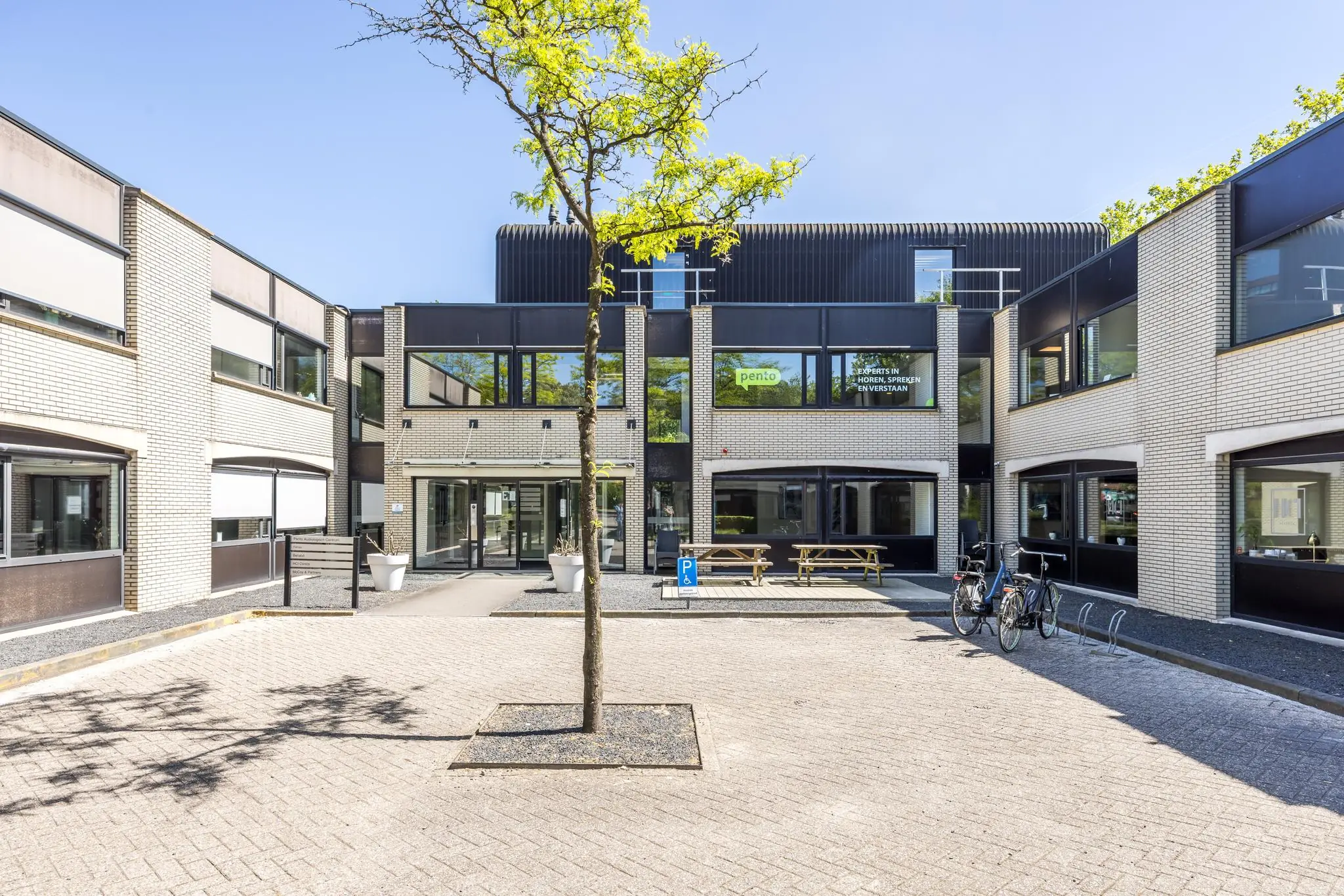 Modern kantoorgebouw aan de Burgemeester Verderlaan met fietsenrek, picknicktafels en boom op de voorgrond.