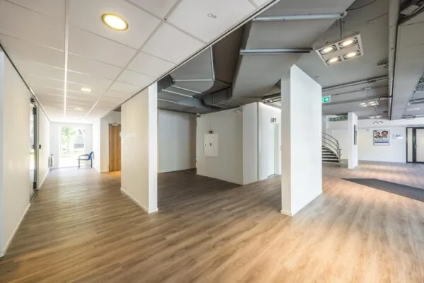 Lichte en moderne entreehal van een kantoorpand aan de Burgemeester Verderlaan met houten vloer, zichtbare ventilatiebuizen en een lift.
