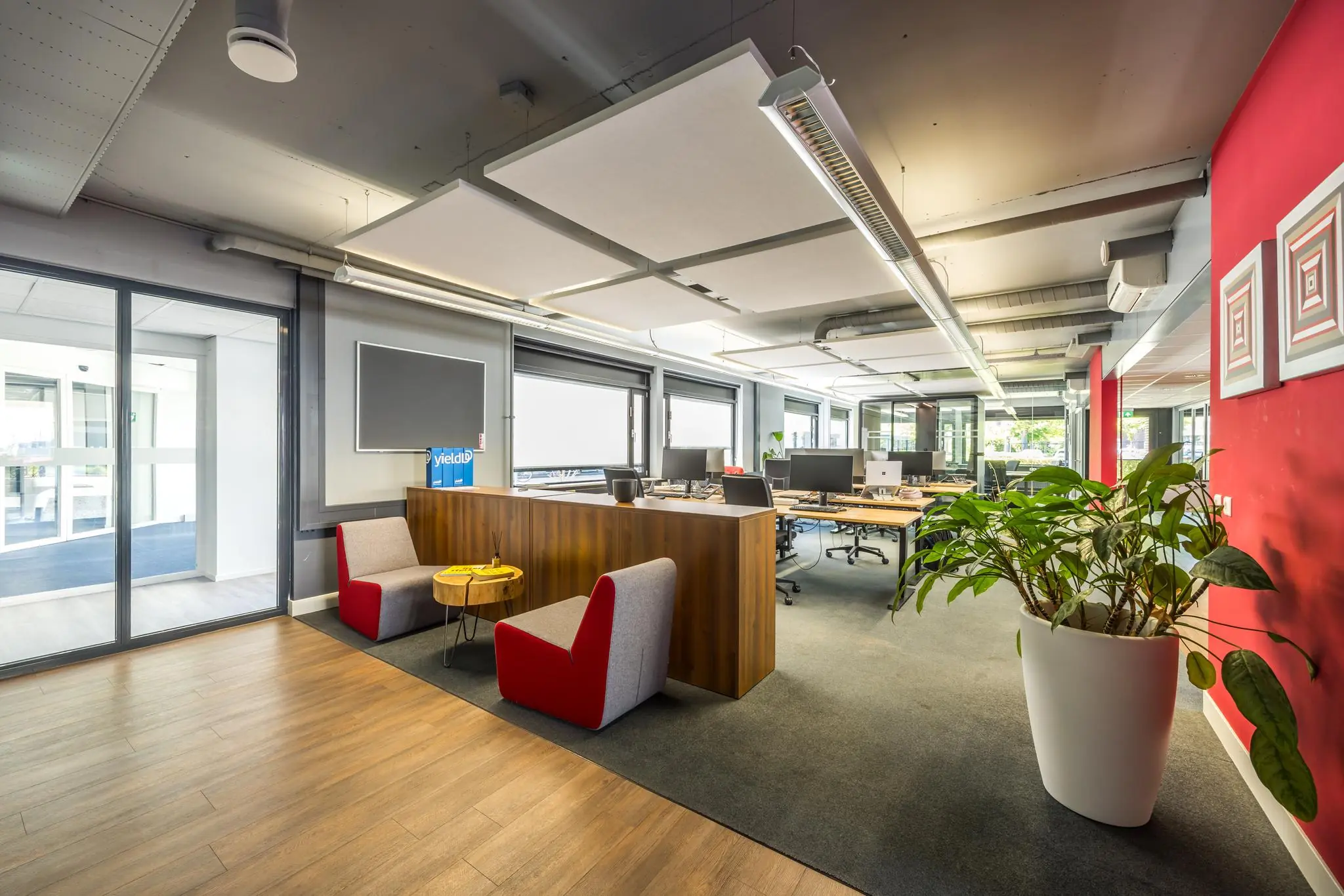 Modern kantoorinterieur met open werkplekken, een zitgedeelte en een grote plant bij een rode accentmuur.