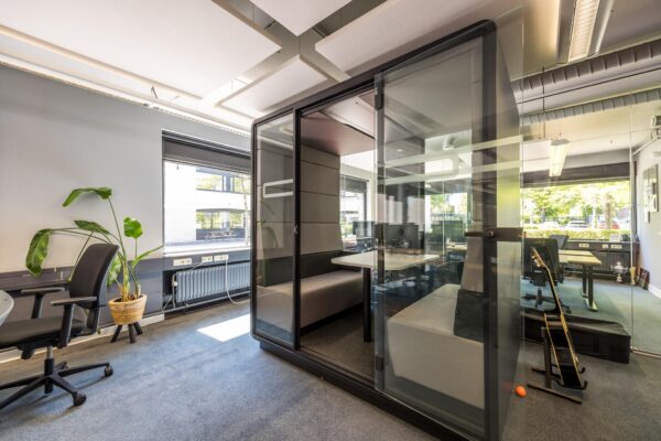 Moderne kantooruimte met een glazen vergaderpod, bureaus, plant en gitaar in een lichte, open omgeving aan de Burgemeester Verderlaan.