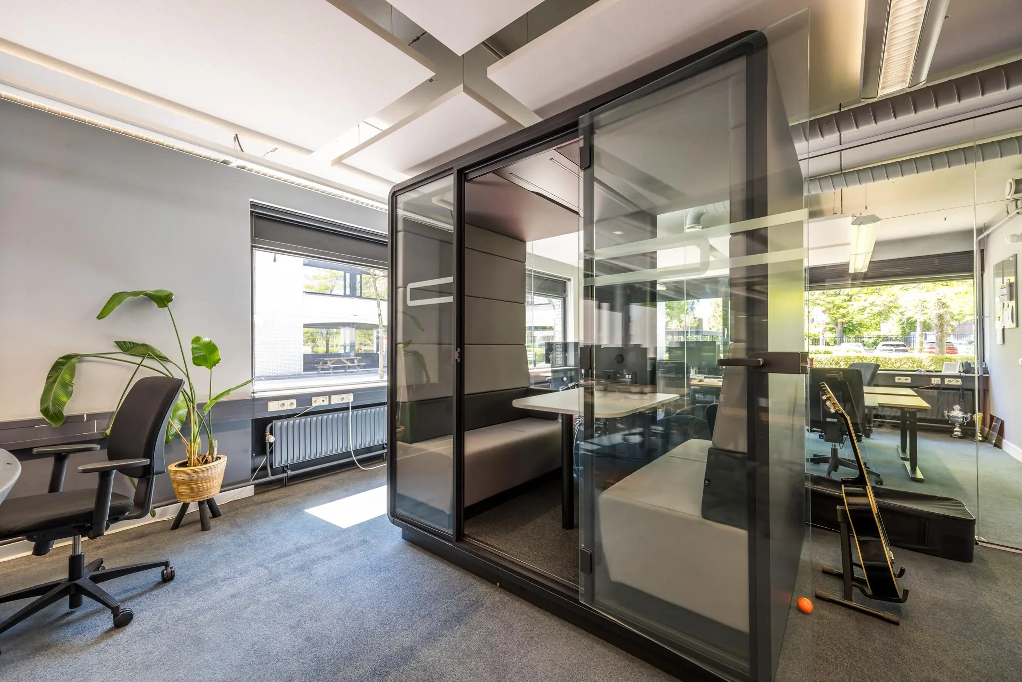 Moderne kantooruimte met een glazen vergaderpod, bureaus, plant en gitaar in een lichte, open omgeving aan de Burgemeester Verderlaan.