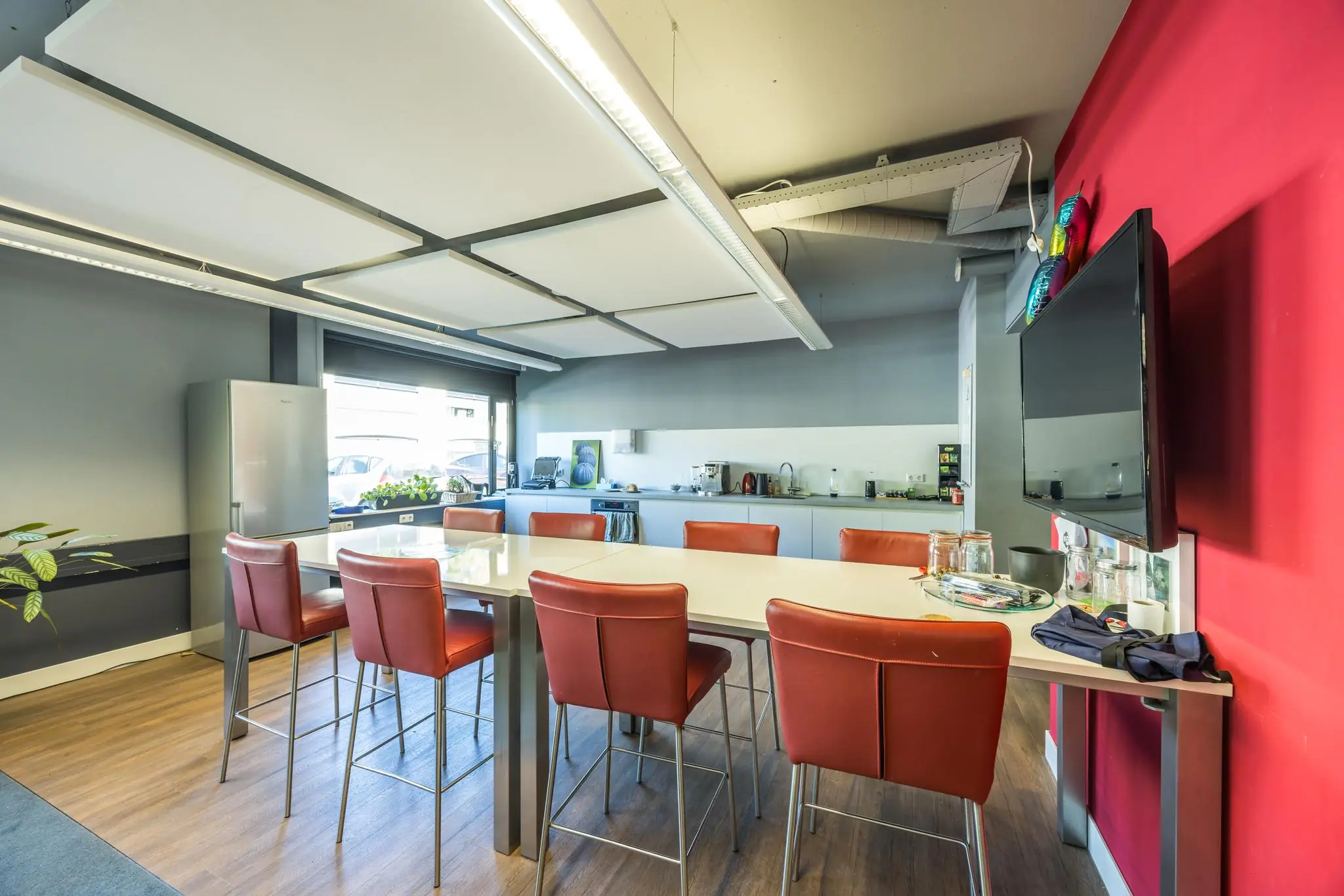 Moderne kantoorkeuken met een grote tafel, rode stoelen, koffiehoek en een flatscreen aan de muur.
