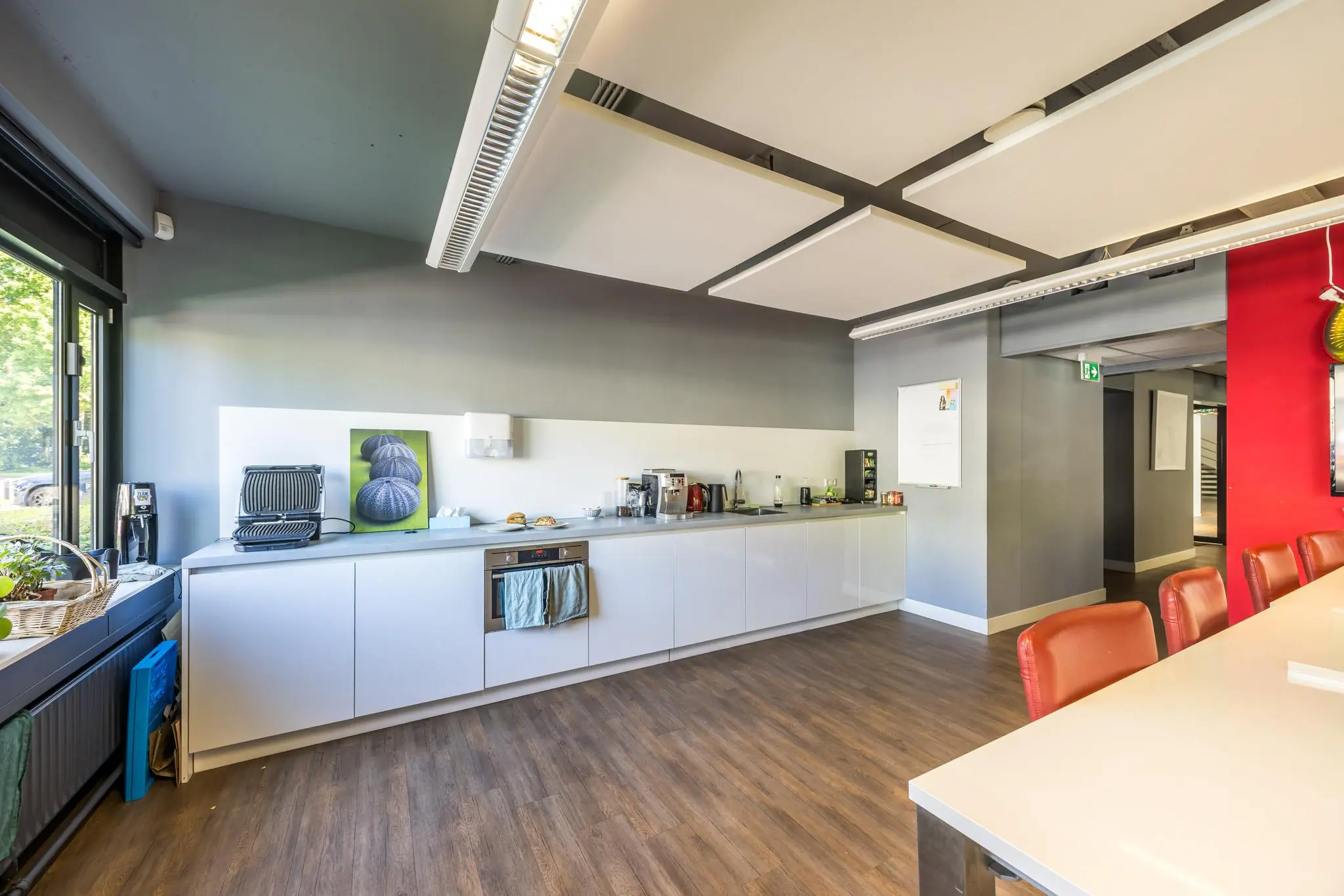 Moderne kantoorkeuken met witte kasten, diverse koffiemachines en een vergadertafel met rode stoelen.