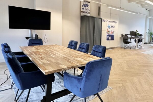 Vergaderruimte met houten tafel, blauwe stoelen, groot scherm en zicht op open kantoorruimte aan de Paulus Potterstraat.