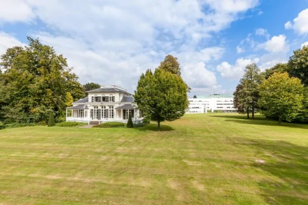 Witte villa Sparrenheuvel met groot gazon en omliggende bomen onder een deels bewolkte hemel.