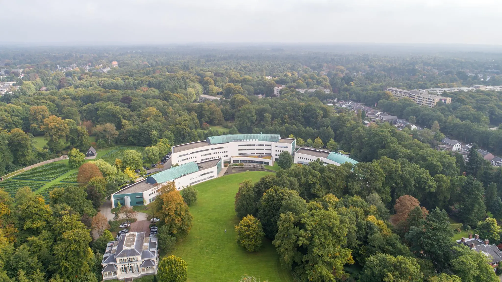 Luchtfoto van het witte, moderne gebouw Sparrenheuvel, omringd door bomen en groen in een bosrijke omgeving.