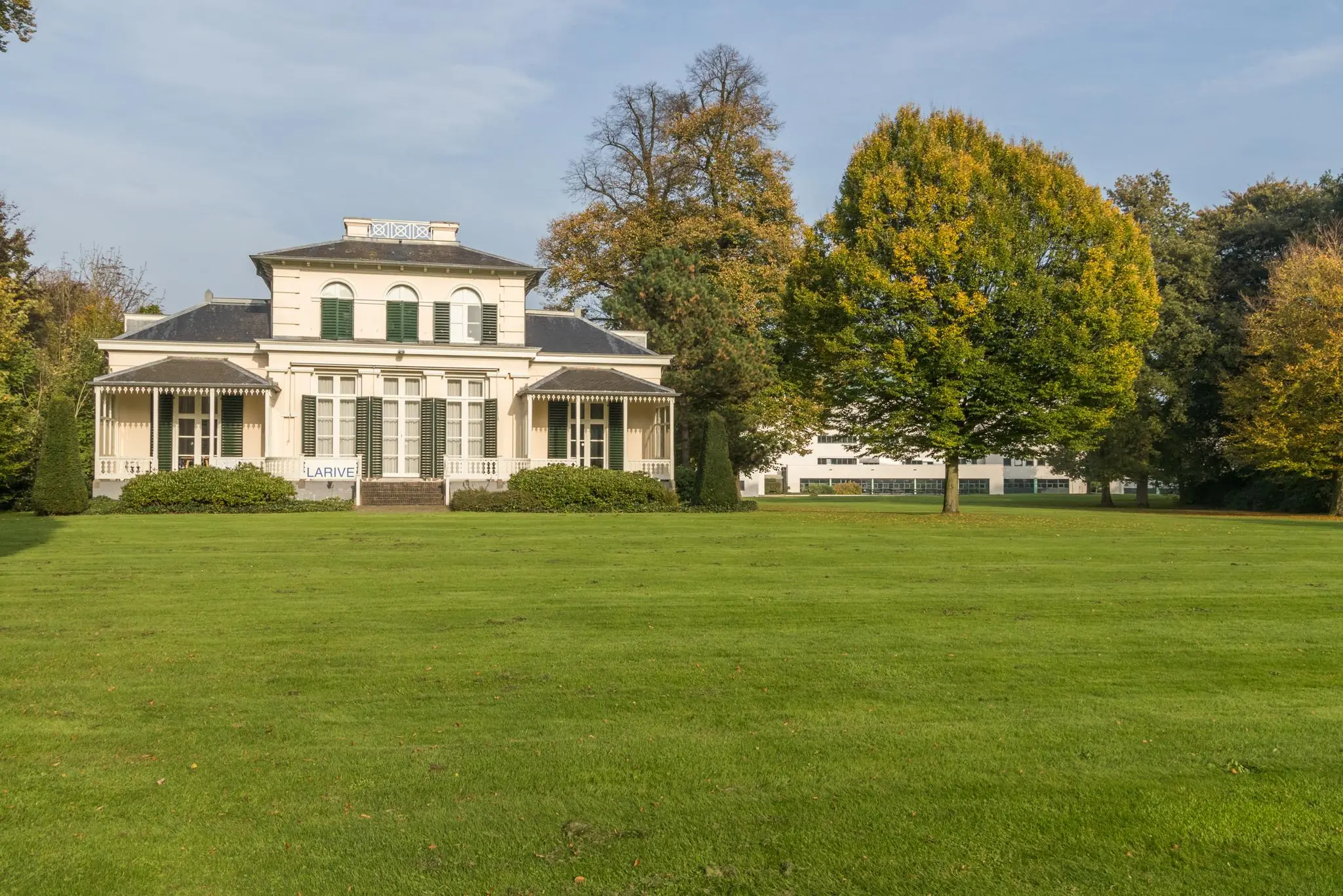 Wit herenhuis Villa Sparrenheuvel met veranda’s en groene luiken, gelegen aan een groot grasveld met bomen.
