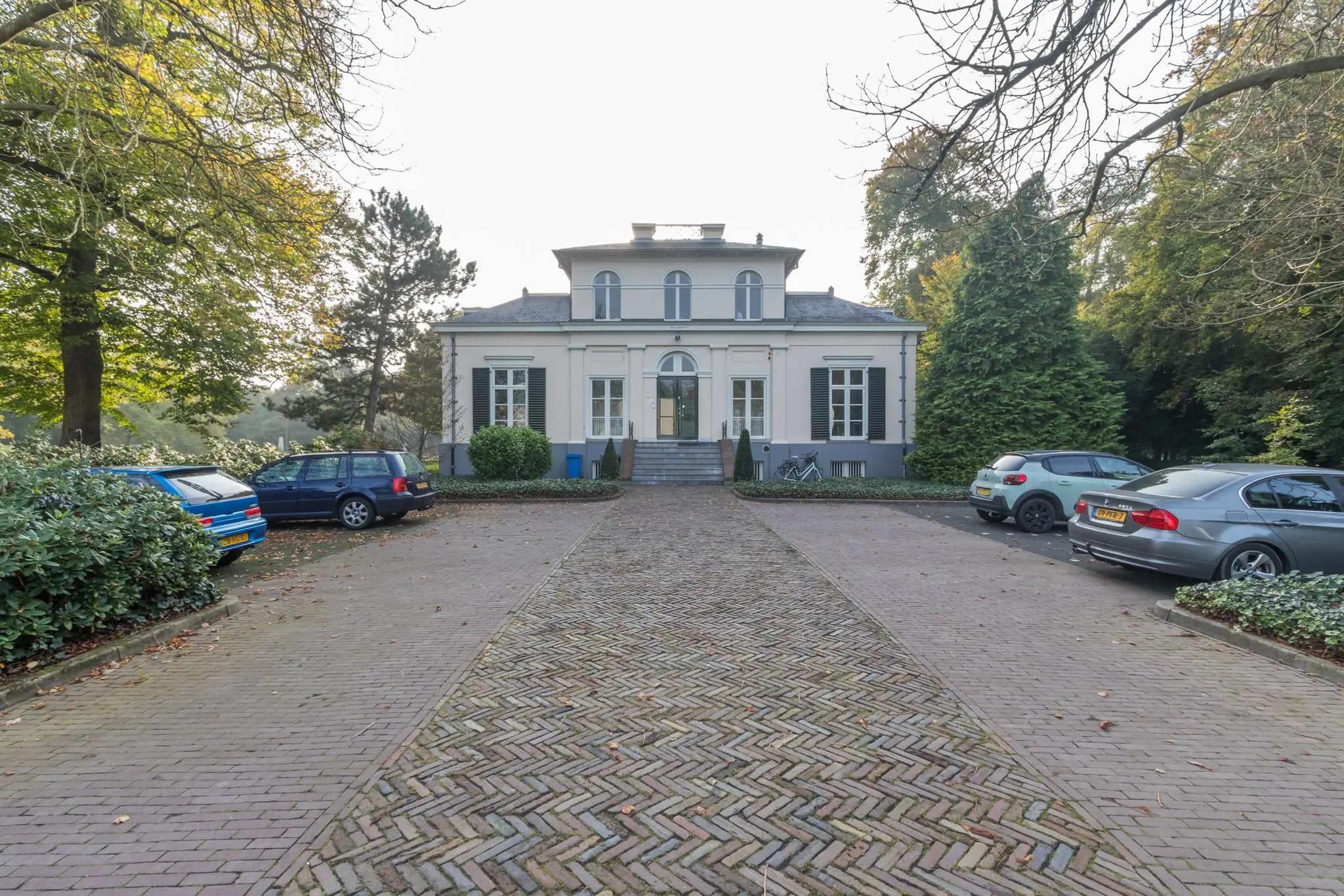 Vooraanzicht van landhuis Sparrenheuvel met oprijlaan en geparkeerde auto's.