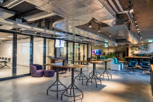 Moderne, industrieel ingerichte lounge- en werkruimte aan de Wibautstraat met hoge tafels, zitplekken en een opvallend plafond met zichtbare luchtkanalen.