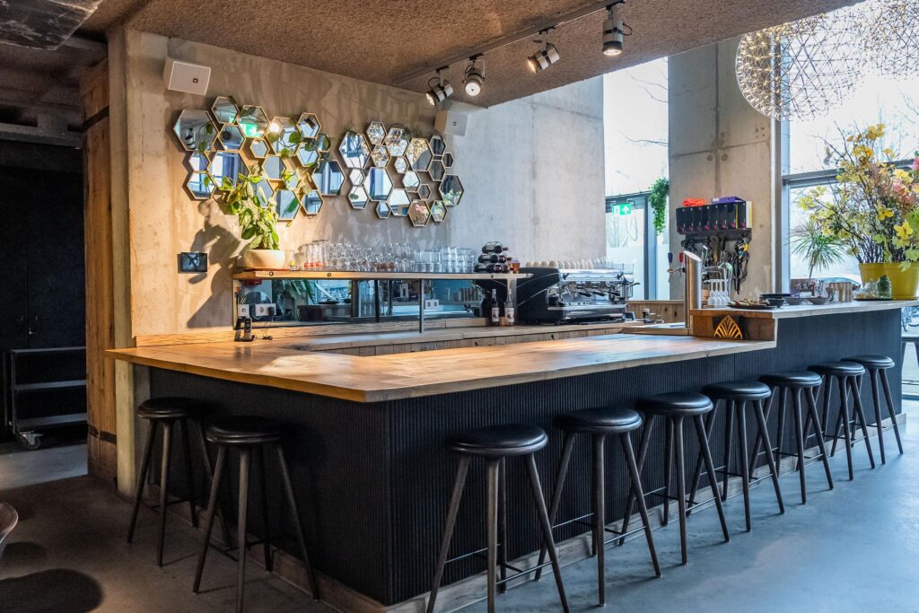 Moderne koffietbar aan de Wibautstraat met houten bar, zwarte barkrukken en decoratieve zeshoekige spiegels aan de muur.