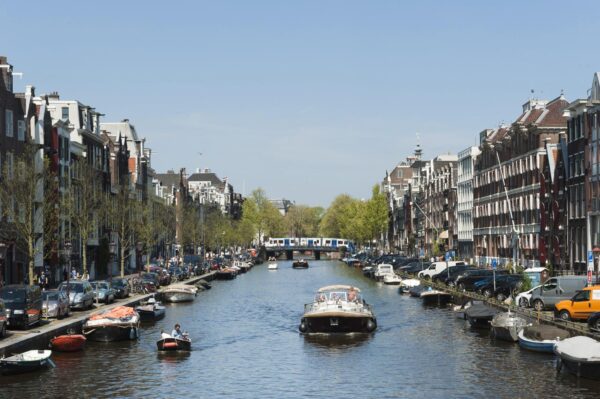 Zicht op de Vijzelgracht in Amsterdam met boten op het water, geparkeerde auto's langs de kant en een metro die over de brug rijdt.