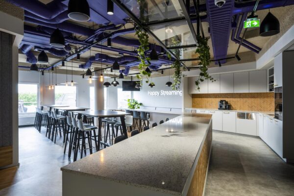 Moderne kantoorkeuken aan de Vijzelgracht met hoge tafels, een lange bar, hangplanten en de tekst "Happy Streaming" op de muur.