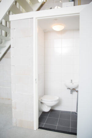 Kleine, witte toiletruimte met hangtoilet en fonteintje onder een trap in een casco bedrijfsruimte aan de Laan van s-Gravenmade.