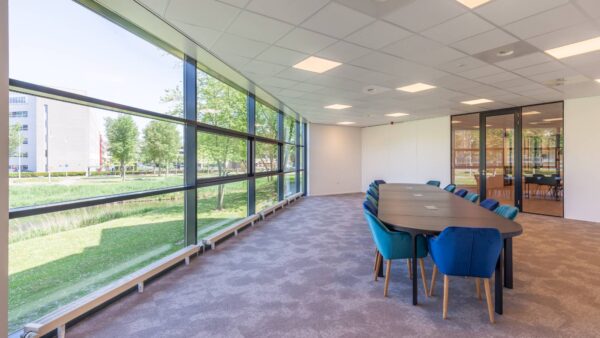 Een lichte vergaderruimte met een lange tafel, blauwe stoelen en grote ramen met uitzicht op buitengroen.