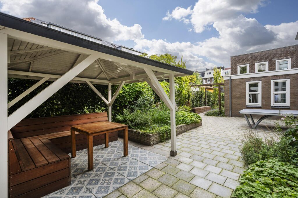 Overdekt zitgedeelte met houten bank en tafel in een groene, betegelde binnentuin aan de Hillegomstraat.