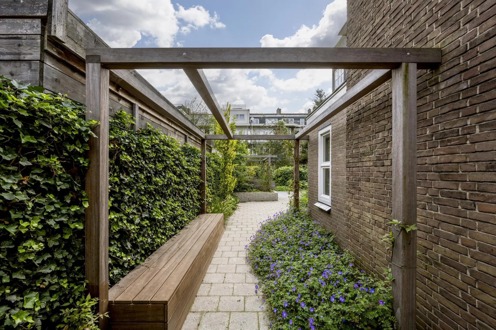 Smalle, betegelde tuin aan de Hillegomstraat met houten pergola, klimplanten en paarse bloemen langs een bakstenen gevel.